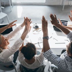 Stylish african student in wristwatch slapping hands with friends, sitting near computer. Portrait from back of joyful office employees fooling around during workday and joking..